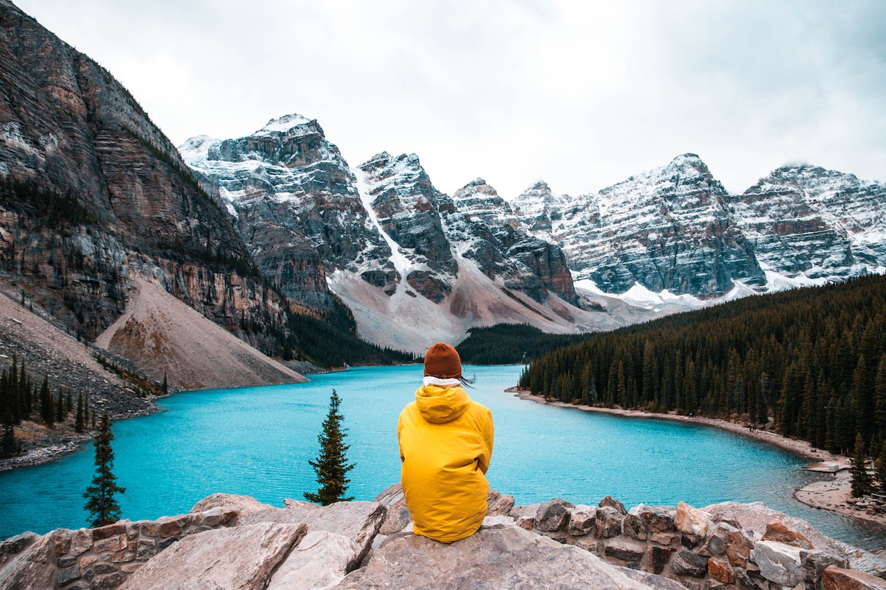 身穿黄色卫衣的人在日间坐在湖边的石头上，远处是被雪覆盖的山